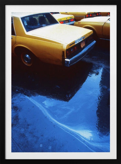 A close-up, angled view of the rear of a yellow taxi cab on a wet street. The car's dark reflection is cast upon a large, vibrant blue puddle in the foreground, with visible ripples from the rain. Wall Art