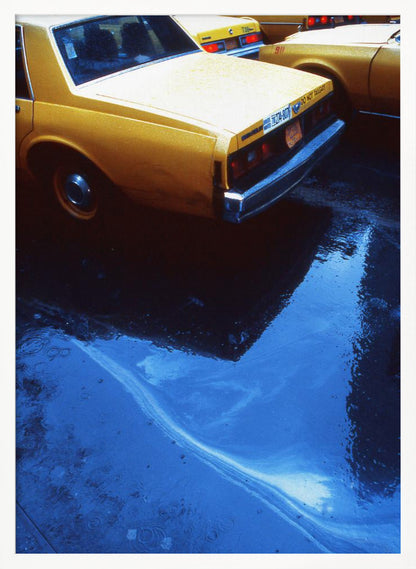 A close-up, angled view of the rear of a yellow taxi cab on a wet street. The car's dark reflection is cast upon a large, vibrant blue puddle in the foreground, with visible ripples from the rain. Wall Art