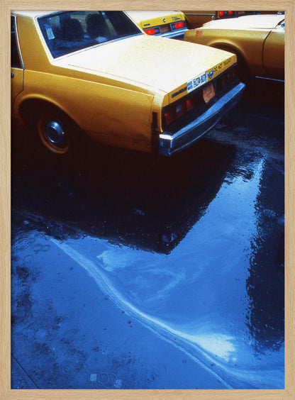 A close-up, angled view of the rear of a yellow taxi cab on a wet street. The car's dark reflection is cast upon a large, vibrant blue puddle in the foreground, with visible ripples from the rain. Wall Art