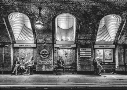 A framed, dramatic black and white photograph of the Baker Street underground station platform in London. Passengers wait on benches beneath iconic brick arches and large grated windows, with signs for 'Baker Street' and the London Underground roundel clearly visible. Print