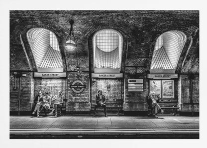 A framed, dramatic black and white photograph of the Baker Street underground station platform in London. Passengers wait on benches beneath iconic brick arches and large grated windows, with signs for 'Baker Street' and the London Underground roundel clearly visible. Print