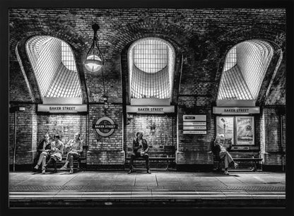 A framed, dramatic black and white photograph of the Baker Street underground station platform in London. Passengers wait on benches beneath iconic brick arches and large grated windows, with signs for 'Baker Street' and the London Underground roundel clearly visible. Print