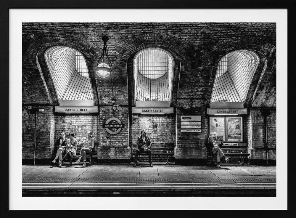 A framed, dramatic black and white photograph of the Baker Street underground station platform in London. Passengers wait on benches beneath iconic brick arches and large grated windows, with signs for 'Baker Street' and the London Underground roundel clearly visible. Print