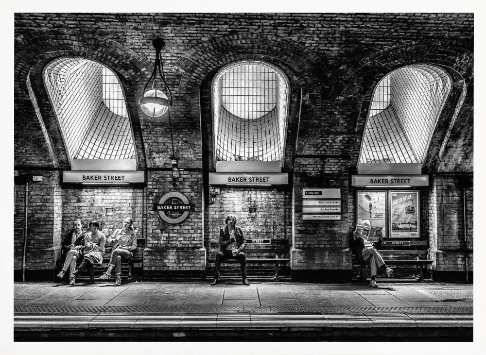 A framed, dramatic black and white photograph of the Baker Street underground station platform in London. Passengers wait on benches beneath iconic brick arches and large grated windows, with signs for 'Baker Street' and the London Underground roundel clearly visible. Print