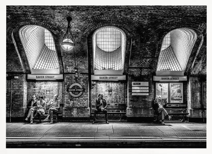 A framed, dramatic black and white photograph of the Baker Street underground station platform in London. Passengers wait on benches beneath iconic brick arches and large grated windows, with signs for 'Baker Street' and the London Underground roundel clearly visible. Print
