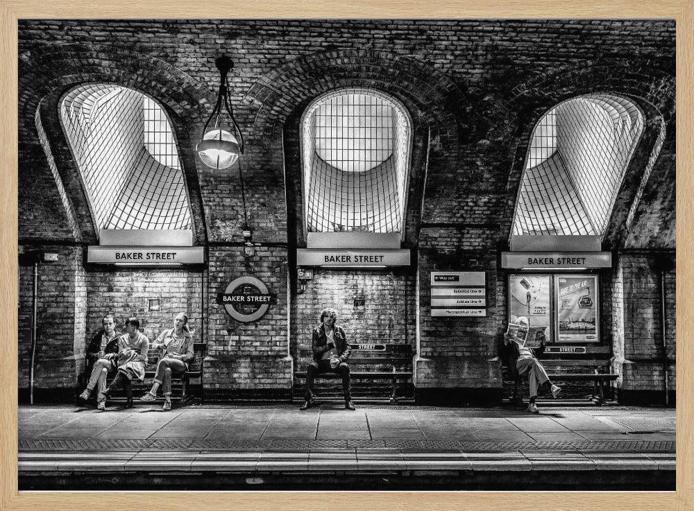 A framed, dramatic black and white photograph of the Baker Street underground station platform in London. Passengers wait on benches beneath iconic brick arches and large grated windows, with signs for 'Baker Street' and the London Underground roundel clearly visible. Print