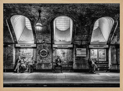 A framed, dramatic black and white photograph of the Baker Street underground station platform in London. Passengers wait on benches beneath iconic brick arches and large grated windows, with signs for 'Baker Street' and the London Underground roundel clearly visible. Print