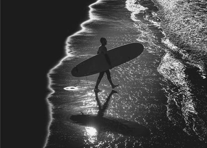 A dramatic, high-contrast, black and white aerial view of a silhouetted surfer carrying their surfboard along the shoreline. The sun glints off the wet sand and the dark water, creating a sparkling effect and a clear reflection of the surfer. Poster