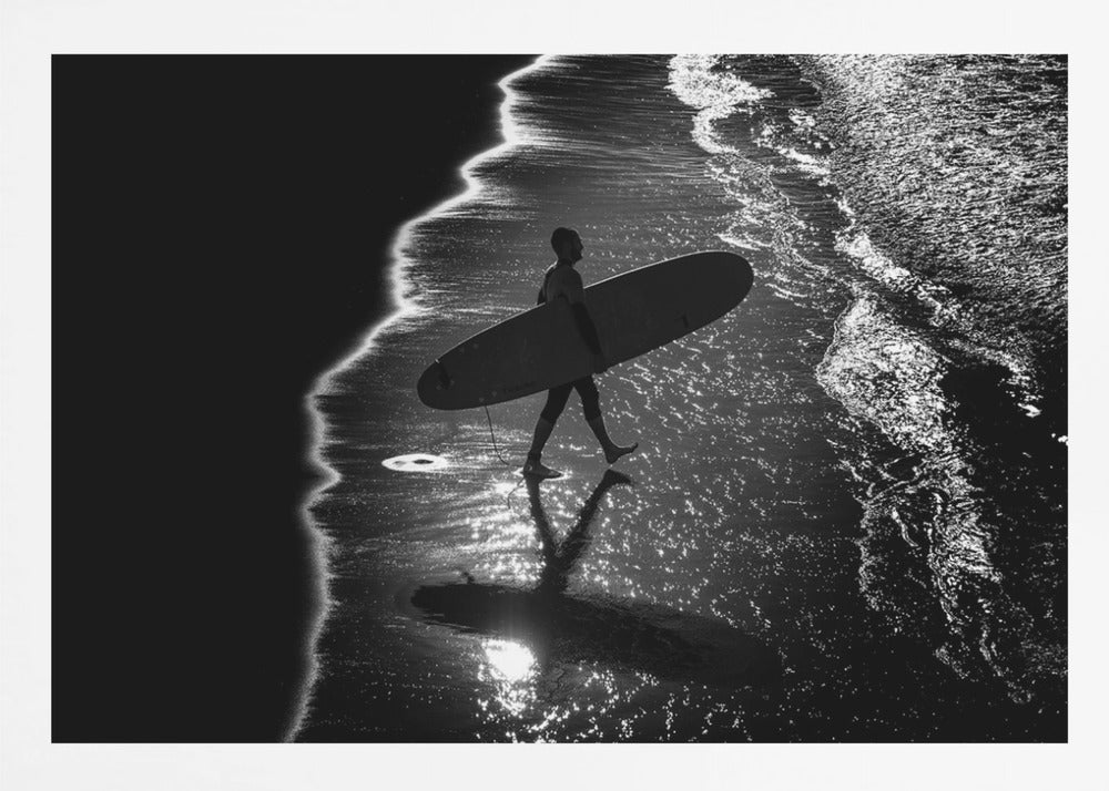 A dramatic, high-contrast, black and white aerial view of a silhouetted surfer carrying their surfboard along the shoreline. The sun glints off the wet sand and the dark water, creating a sparkling effect and a clear reflection of the surfer. Poster