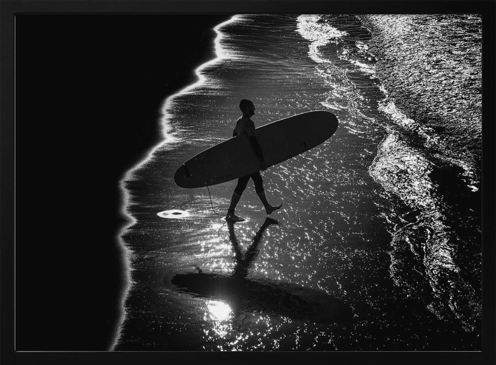 A dramatic, high-contrast, black and white aerial view of a silhouetted surfer carrying their surfboard along the shoreline. The sun glints off the wet sand and the dark water, creating a sparkling effect and a clear reflection of the surfer. Poster