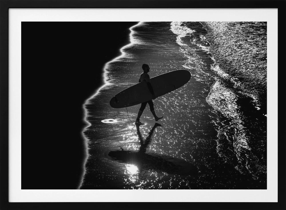 A dramatic, high-contrast, black and white aerial view of a silhouetted surfer carrying their surfboard along the shoreline. The sun glints off the wet sand and the dark water, creating a sparkling effect and a clear reflection of the surfer. Poster