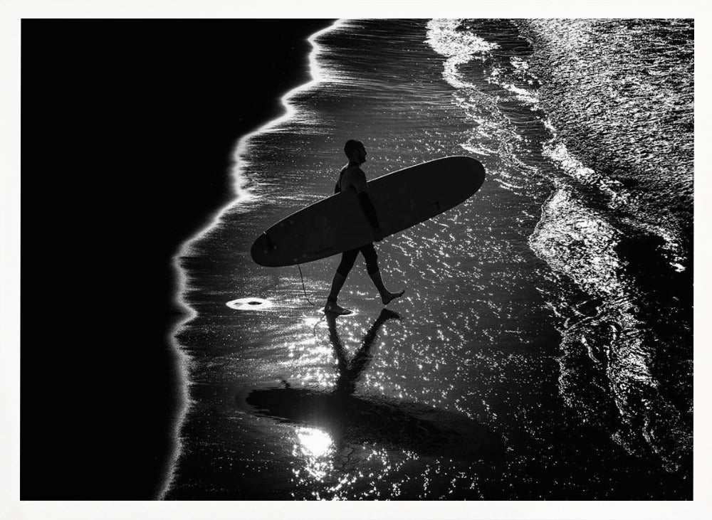 A dramatic, high-contrast, black and white aerial view of a silhouetted surfer carrying their surfboard along the shoreline. The sun glints off the wet sand and the dark water, creating a sparkling effect and a clear reflection of the surfer. Poster