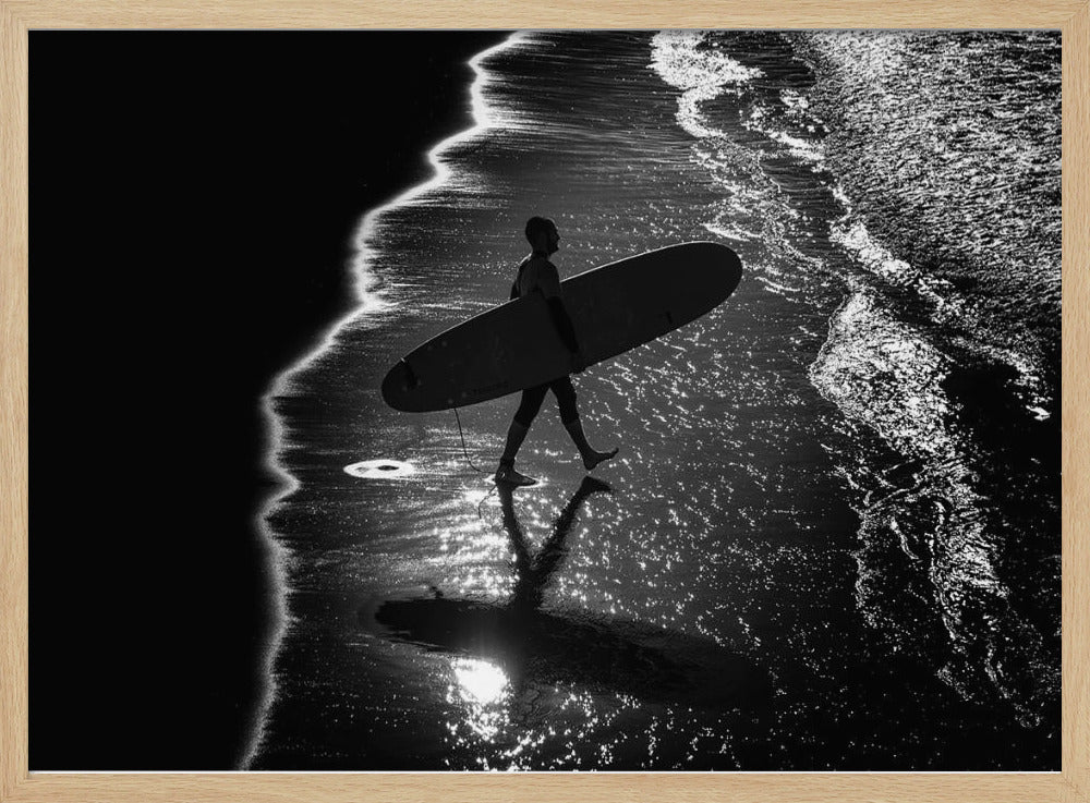 A dramatic, high-contrast, black and white aerial view of a silhouetted surfer carrying their surfboard along the shoreline. The sun glints off the wet sand and the dark water, creating a sparkling effect and a clear reflection of the surfer. Poster