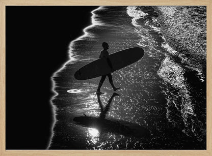 A dramatic, high-contrast, black and white aerial view of a silhouetted surfer carrying their surfboard along the shoreline. The sun glints off the wet sand and the dark water, creating a sparkling effect and a clear reflection of the surfer. Poster