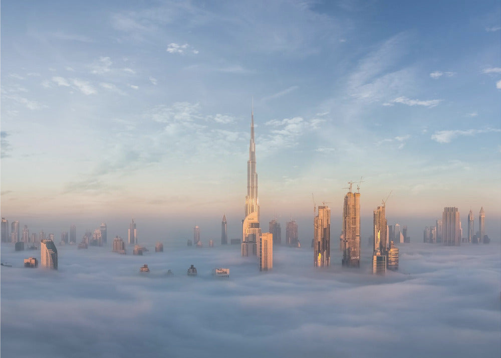 A framed photograph of the Dubai skyline, with the Burj Khalifa and other skyscrapers piercing through a thick blanket of low-lying clouds or fog. The scene is bathed in the warm, golden light of sunrise or sunset against a pale blue sky. Poster