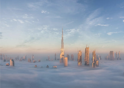 A framed photograph of the Dubai skyline, with the Burj Khalifa and other skyscrapers piercing through a thick blanket of low-lying clouds or fog. The scene is bathed in the warm, golden light of sunrise or sunset against a pale blue sky. Poster