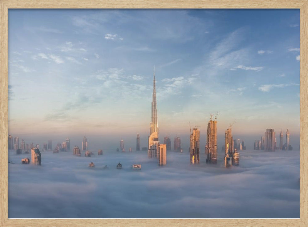 A framed photograph of the Dubai skyline, with the Burj Khalifa and other skyscrapers piercing through a thick blanket of low-lying clouds or fog. The scene is bathed in the warm, golden light of sunrise or sunset against a pale blue sky. Poster