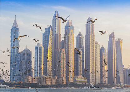 A framed artwork of the Dubai city skyline, featuring modern skyscrapers bathed in the golden light of sunset. A flock of seagulls flies in the foreground above the water, where several yachts are docked. Artwork