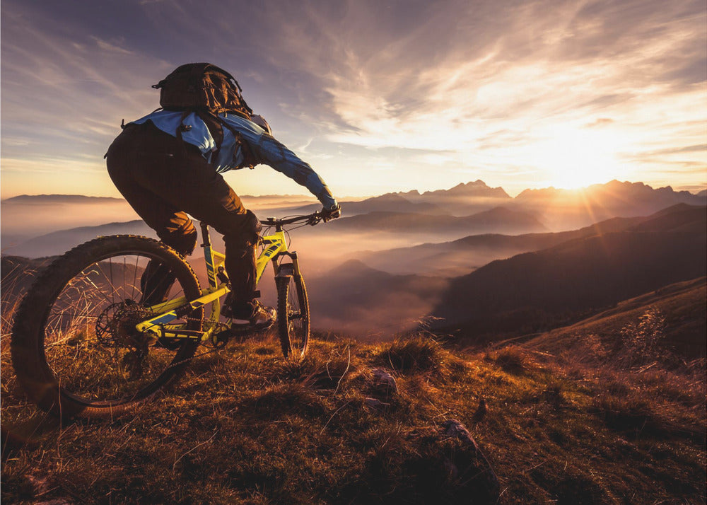 A mountain biker, seen from behind, stops at the top of a grassy hill to admire a spectacular sunset over a vast mountain range. The valleys below are filled with mist, and the sun's golden rays create a stunning starburst effect against the silhouetted peaks. Print
