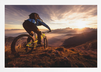 A mountain biker, seen from behind, stops at the top of a grassy hill to admire a spectacular sunset over a vast mountain range. The valleys below are filled with mist, and the sun's golden rays create a stunning starburst effect against the silhouetted peaks. Print