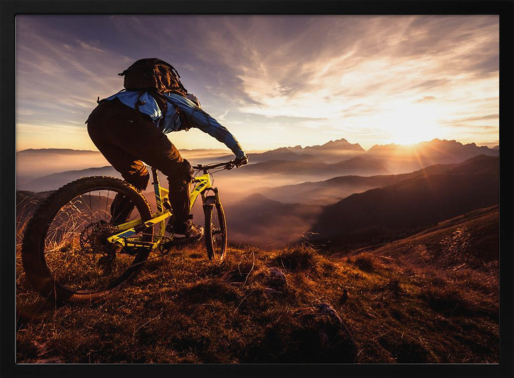 A mountain biker, seen from behind, stops at the top of a grassy hill to admire a spectacular sunset over a vast mountain range. The valleys below are filled with mist, and the sun's golden rays create a stunning starburst effect against the silhouetted peaks. Print