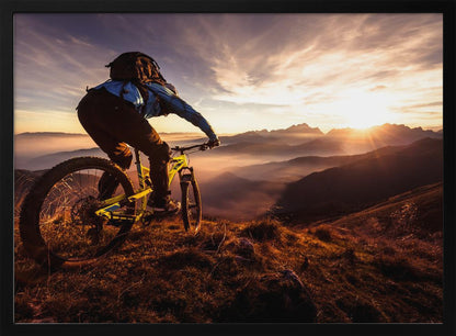 A mountain biker, seen from behind, stops at the top of a grassy hill to admire a spectacular sunset over a vast mountain range. The valleys below are filled with mist, and the sun's golden rays create a stunning starburst effect against the silhouetted peaks. Print