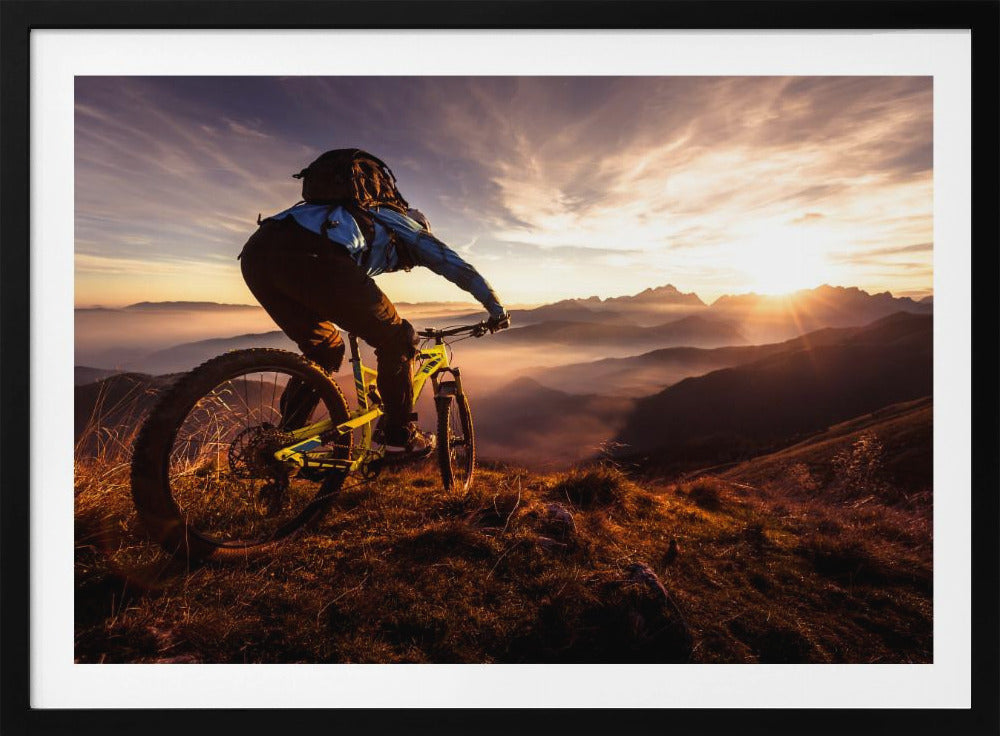 A mountain biker, seen from behind, stops at the top of a grassy hill to admire a spectacular sunset over a vast mountain range. The valleys below are filled with mist, and the sun's golden rays create a stunning starburst effect against the silhouetted peaks. Print