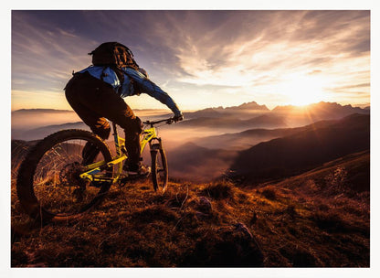 A mountain biker, seen from behind, stops at the top of a grassy hill to admire a spectacular sunset over a vast mountain range. The valleys below are filled with mist, and the sun's golden rays create a stunning starburst effect against the silhouetted peaks. Print