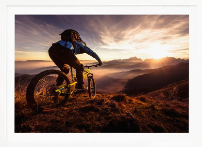 A mountain biker, seen from behind, stops at the top of a grassy hill to admire a spectacular sunset over a vast mountain range. The valleys below are filled with mist, and the sun's golden rays create a stunning starburst effect against the silhouetted peaks. Print