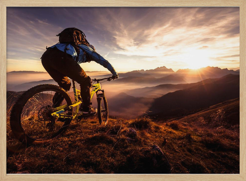 A mountain biker, seen from behind, stops at the top of a grassy hill to admire a spectacular sunset over a vast mountain range. The valleys below are filled with mist, and the sun's golden rays create a stunning starburst effect against the silhouetted peaks. Print