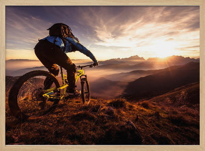 A mountain biker, seen from behind, stops at the top of a grassy hill to admire a spectacular sunset over a vast mountain range. The valleys below are filled with mist, and the sun's golden rays create a stunning starburst effect against the silhouetted peaks. Print