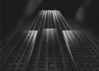 A dramatic, low-angle black and white photograph of the Empire State Building at night. The perspective looks up from the base, emphasizing the skyscraper's height and Art Deco details. Spotlights highlight the vertical lines of the building against a dark, moody sky, all enclosed in a silver frame. Poster
