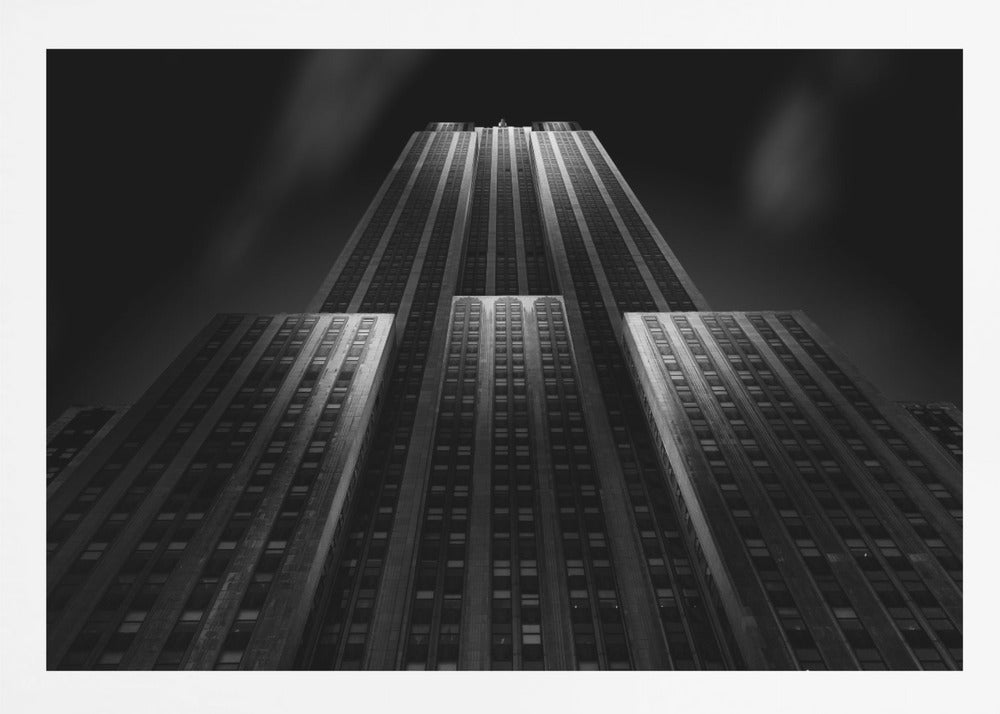 A dramatic, low-angle black and white photograph of the Empire State Building at night. The perspective looks up from the base, emphasizing the skyscraper's height and Art Deco details. Spotlights highlight the vertical lines of the building against a dark, moody sky, all enclosed in a silver frame. Poster