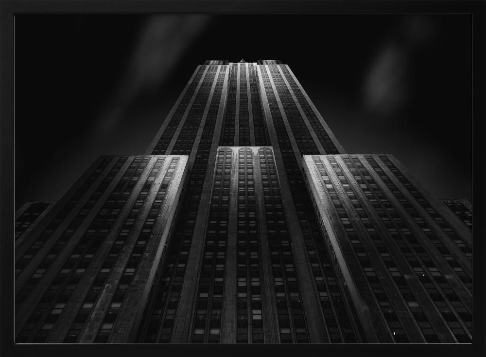 A dramatic, low-angle black and white photograph of the Empire State Building at night. The perspective looks up from the base, emphasizing the skyscraper's height and Art Deco details. Spotlights highlight the vertical lines of the building against a dark, moody sky, all enclosed in a silver frame. Poster
