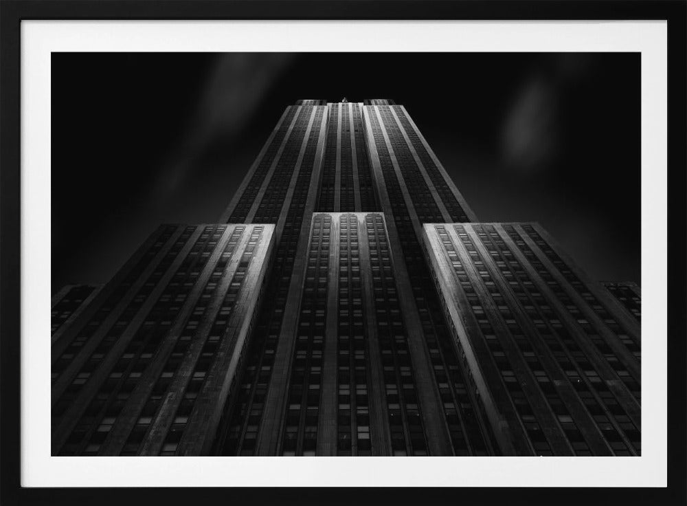 A dramatic, low-angle black and white photograph of the Empire State Building at night. The perspective looks up from the base, emphasizing the skyscraper's height and Art Deco details. Spotlights highlight the vertical lines of the building against a dark, moody sky, all enclosed in a silver frame. Poster