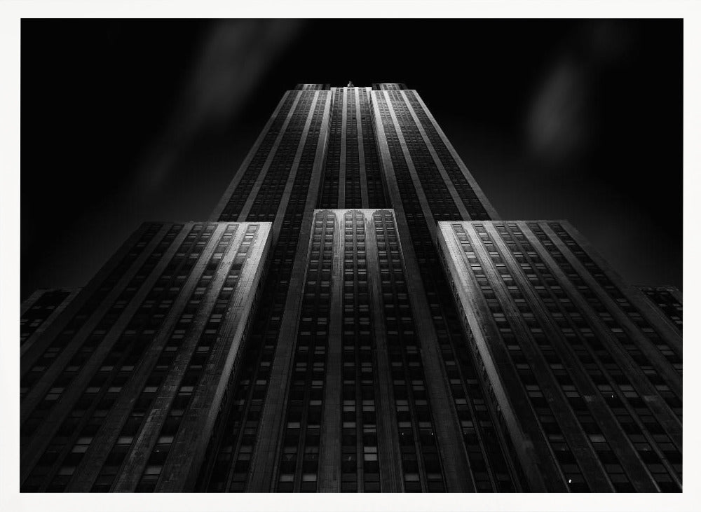 A dramatic, low-angle black and white photograph of the Empire State Building at night. The perspective looks up from the base, emphasizing the skyscraper's height and Art Deco details. Spotlights highlight the vertical lines of the building against a dark, moody sky, all enclosed in a silver frame. Poster