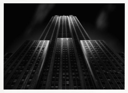 A dramatic, low-angle black and white photograph of the Empire State Building at night. The perspective looks up from the base, emphasizing the skyscraper's height and Art Deco details. Spotlights highlight the vertical lines of the building against a dark, moody sky, all enclosed in a silver frame. Poster