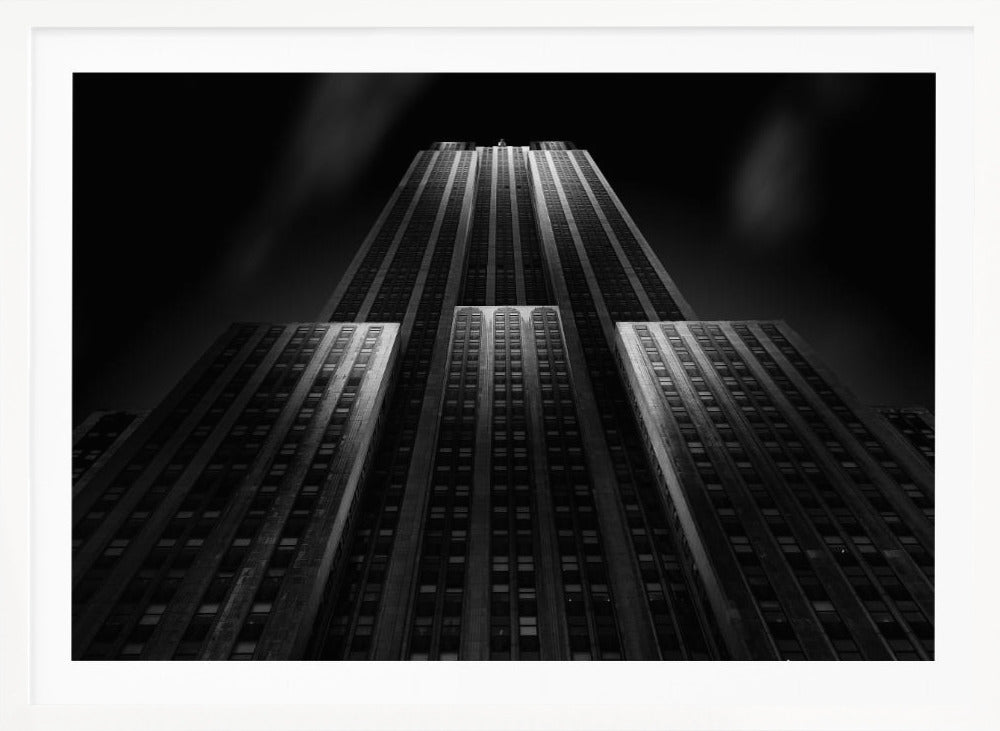 A dramatic, low-angle black and white photograph of the Empire State Building at night. The perspective looks up from the base, emphasizing the skyscraper's height and Art Deco details. Spotlights highlight the vertical lines of the building against a dark, moody sky, all enclosed in a silver frame. Poster