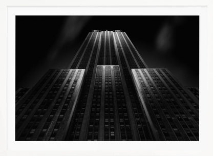 A dramatic, low-angle black and white photograph of the Empire State Building at night. The perspective looks up from the base, emphasizing the skyscraper's height and Art Deco details. Spotlights highlight the vertical lines of the building against a dark, moody sky, all enclosed in a silver frame. Poster