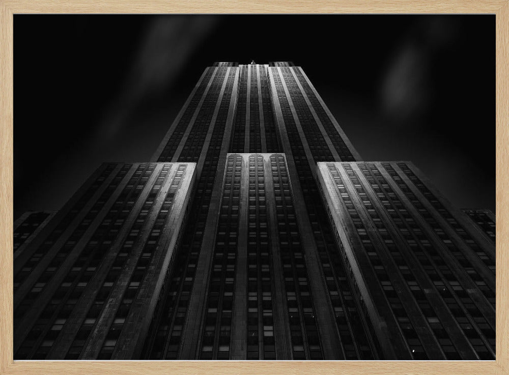 A dramatic, low-angle black and white photograph of the Empire State Building at night. The perspective looks up from the base, emphasizing the skyscraper's height and Art Deco details. Spotlights highlight the vertical lines of the building against a dark, moody sky, all enclosed in a silver frame. Poster