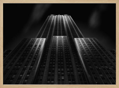 A dramatic, low-angle black and white photograph of the Empire State Building at night. The perspective looks up from the base, emphasizing the skyscraper's height and Art Deco details. Spotlights highlight the vertical lines of the building against a dark, moody sky, all enclosed in a silver frame. Poster