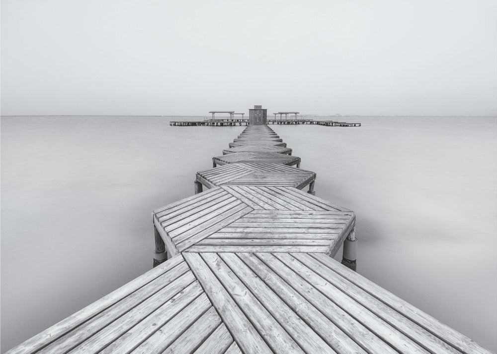 A framed, black and white photograph capturing a long wooden pier from a first-person perspective. The pier, constructed from a series of hexagonal platforms, stretches out into a calm, placid body of water that disappears into a foggy horizon, creating a serene and minimalist scene. Print
