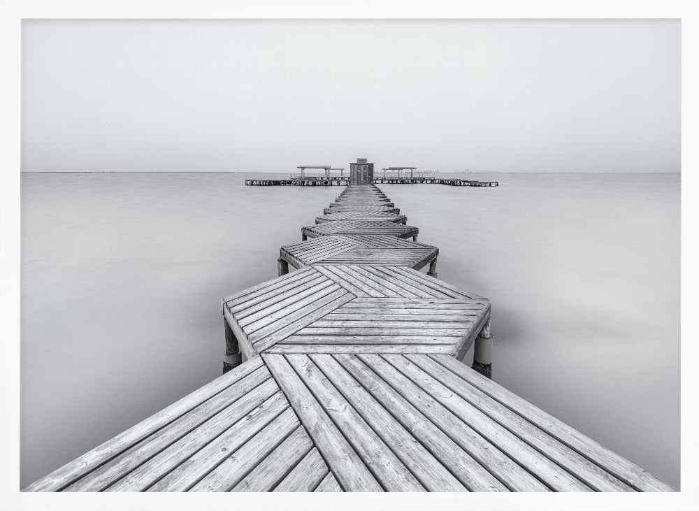 A framed, black and white photograph capturing a long wooden pier from a first-person perspective. The pier, constructed from a series of hexagonal platforms, stretches out into a calm, placid body of water that disappears into a foggy horizon, creating a serene and minimalist scene. Print