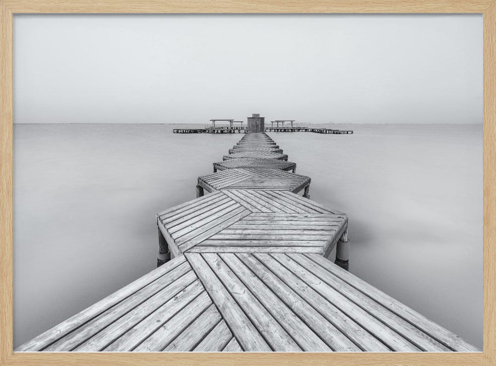 A framed, black and white photograph capturing a long wooden pier from a first-person perspective. The pier, constructed from a series of hexagonal platforms, stretches out into a calm, placid body of water that disappears into a foggy horizon, creating a serene and minimalist scene. Print