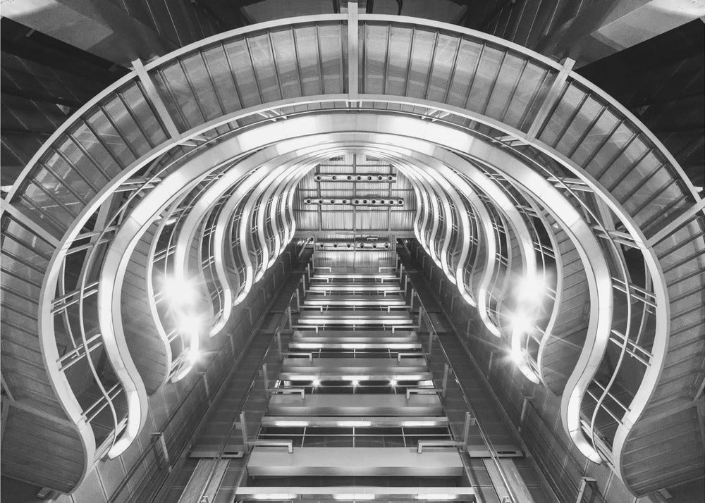 A symmetrical, low-angle black and white photograph of a futuristic escalator or grand staircase. The structure features illuminated, repeating curved arches on both sides, creating a tunnel-like perspective leading upwards. The image is presented within a silver-colored frame. Artwork
