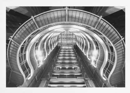 A symmetrical, low-angle black and white photograph of a futuristic escalator or grand staircase. The structure features illuminated, repeating curved arches on both sides, creating a tunnel-like perspective leading upwards. The image is presented within a silver-colored frame. Artwork