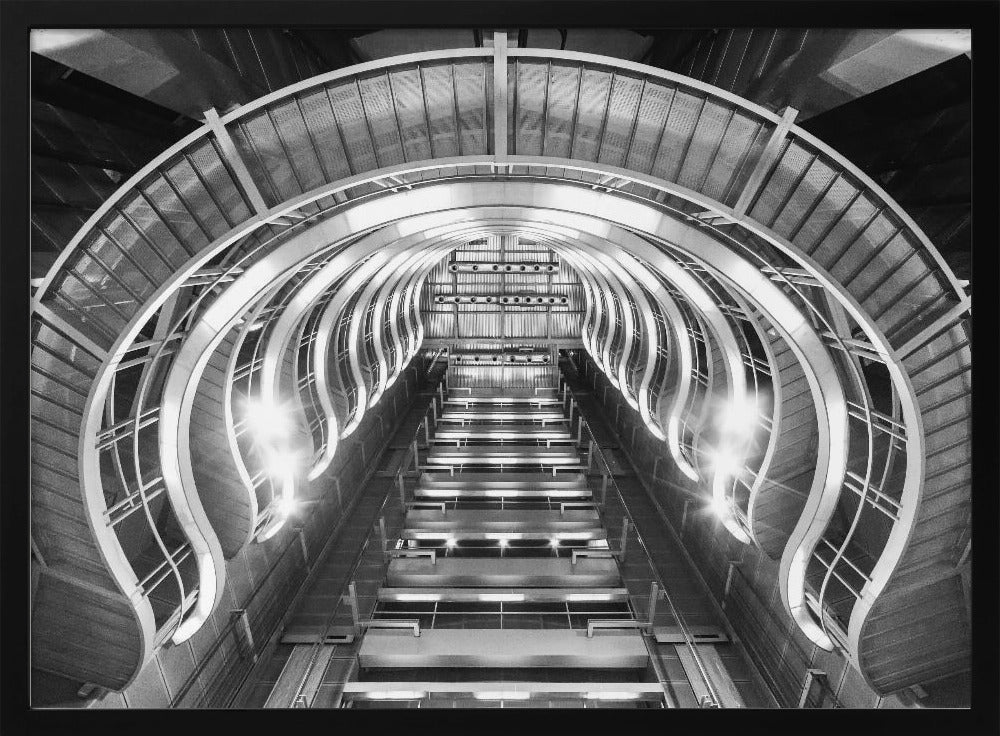 A symmetrical, low-angle black and white photograph of a futuristic escalator or grand staircase. The structure features illuminated, repeating curved arches on both sides, creating a tunnel-like perspective leading upwards. The image is presented within a silver-colored frame. Artwork