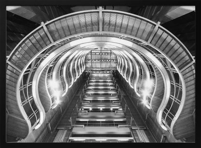 A symmetrical, low-angle black and white photograph of a futuristic escalator or grand staircase. The structure features illuminated, repeating curved arches on both sides, creating a tunnel-like perspective leading upwards. The image is presented within a silver-colored frame. Artwork
