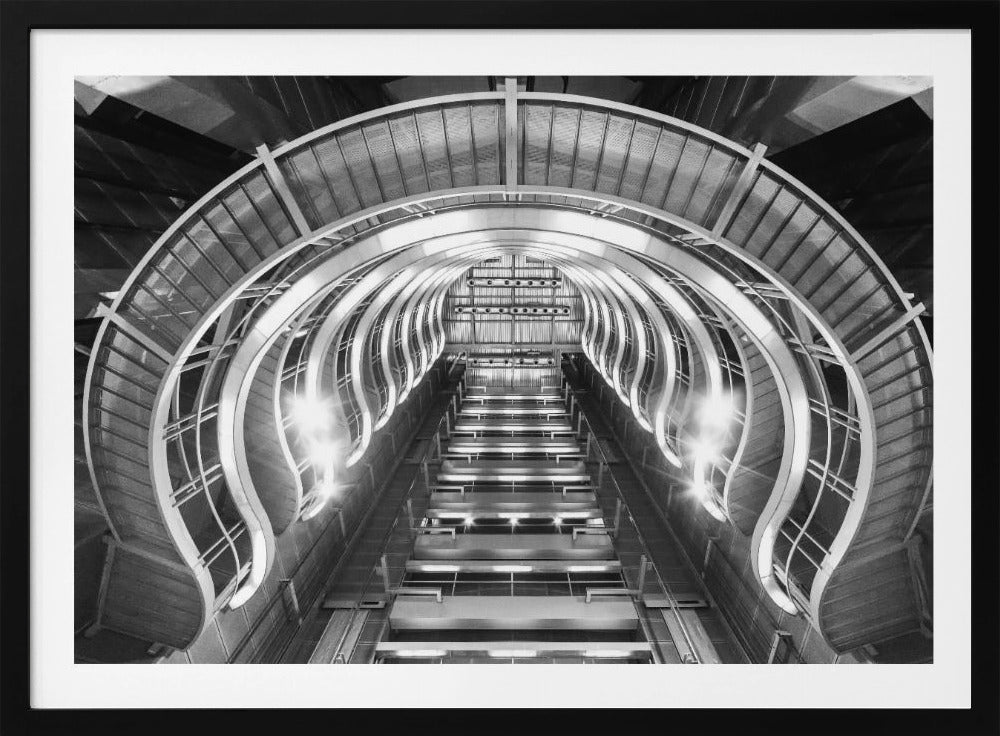A symmetrical, low-angle black and white photograph of a futuristic escalator or grand staircase. The structure features illuminated, repeating curved arches on both sides, creating a tunnel-like perspective leading upwards. The image is presented within a silver-colored frame. Artwork