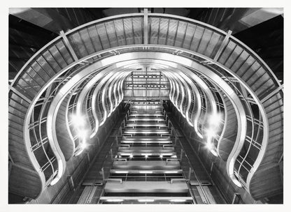 A symmetrical, low-angle black and white photograph of a futuristic escalator or grand staircase. The structure features illuminated, repeating curved arches on both sides, creating a tunnel-like perspective leading upwards. The image is presented within a silver-colored frame. Artwork